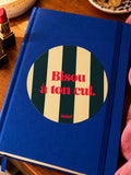 Sticker Muisti Bisou à ton cul collé sur un carnet bleu sur une table de nuit avec rouge à lèvres