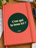 Sticker Muisti C’est qui la boss ici ? collé sur un carnet corail sur un bureau