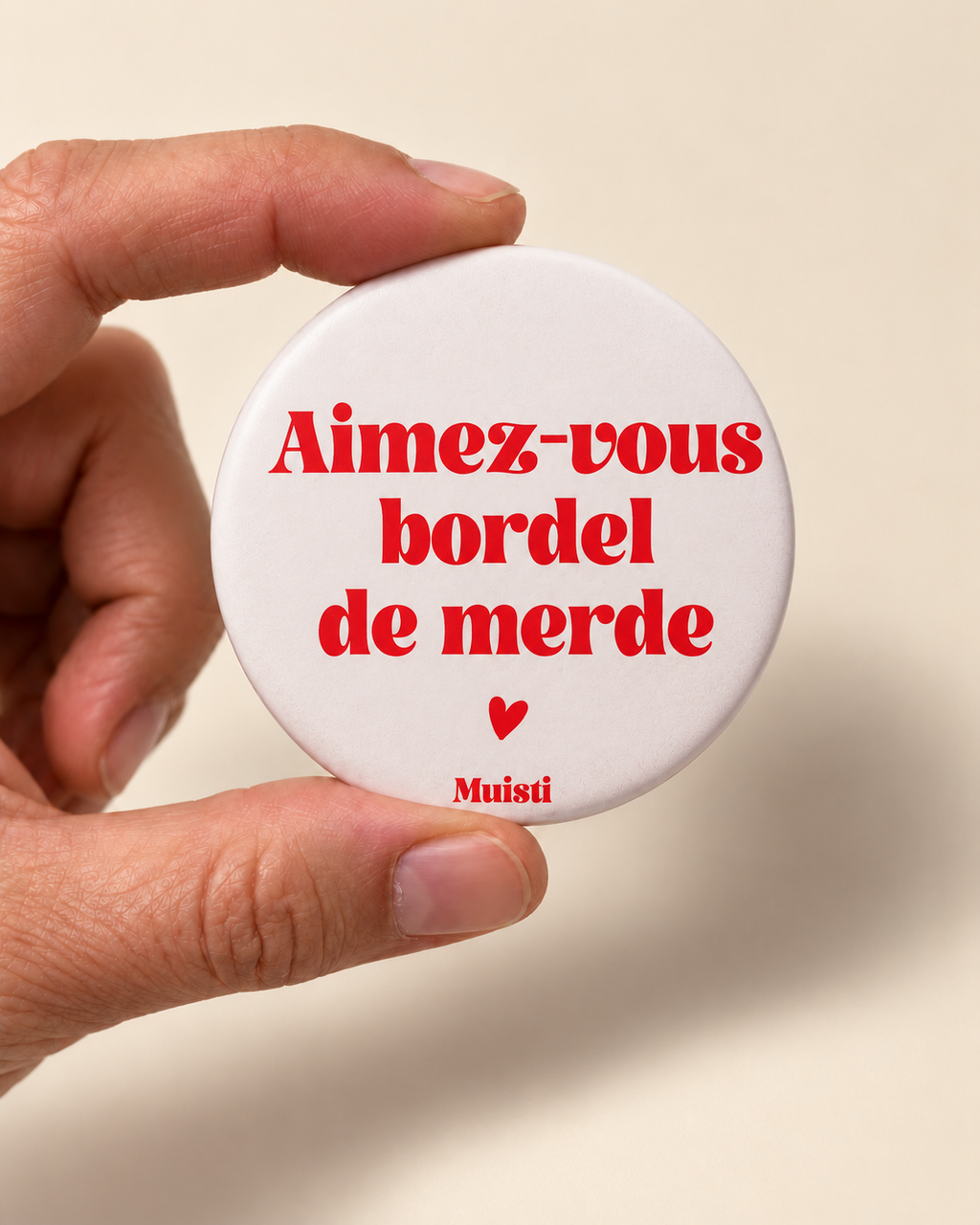 Magnet Muisti « Aimez-vous bordel de merde » tenu entre le pouce et l’index sur fond ivoire.
