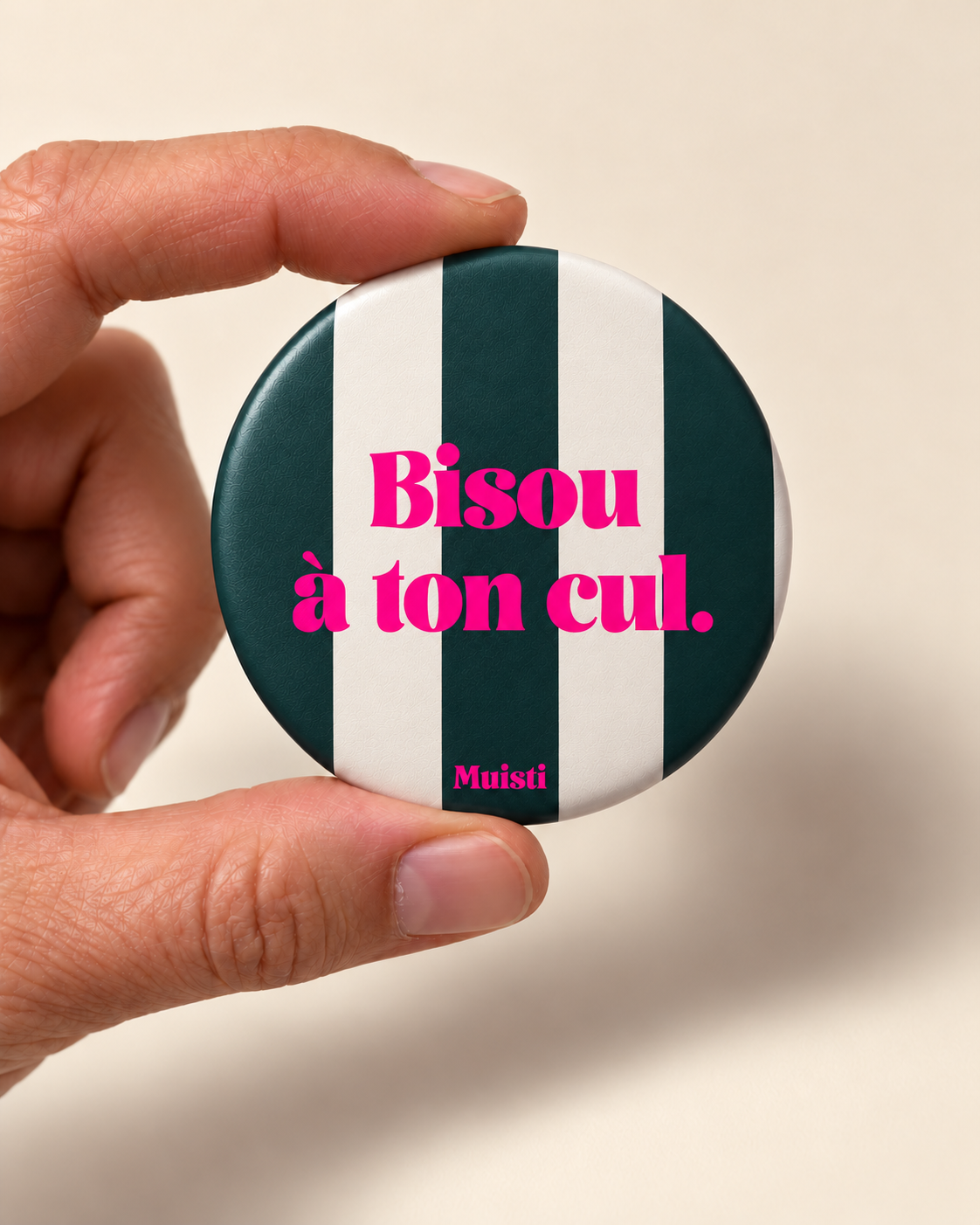 Magnet Muisti « Bisou à ton cul » tenu entre le pouce et l’index sur fond ivoire.