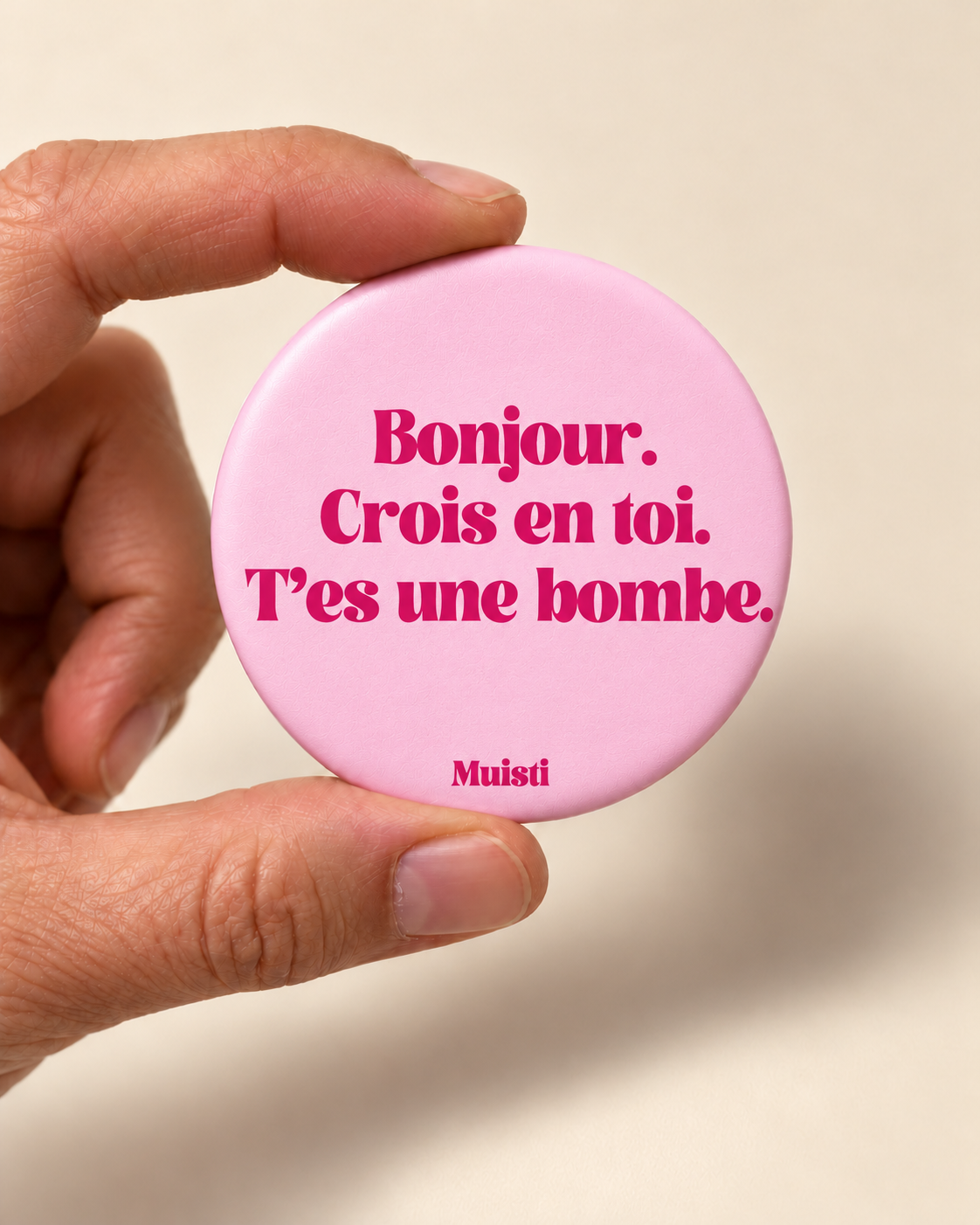 Magnet Muisti « Bonjour. Crois en toi. T’es une bombe. » tenu entre le pouce et l’index sur fond ivoire.