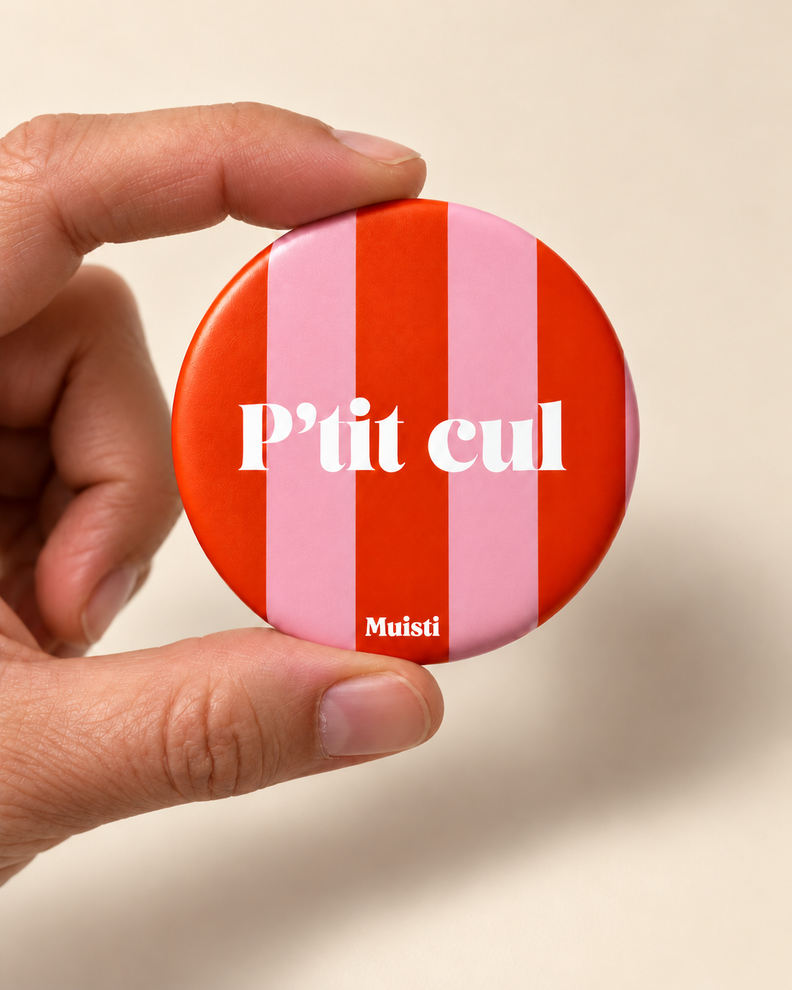 Magnet Muisti « P’tit cul » tenu entre le pouce et l’index sur fond ivoire.