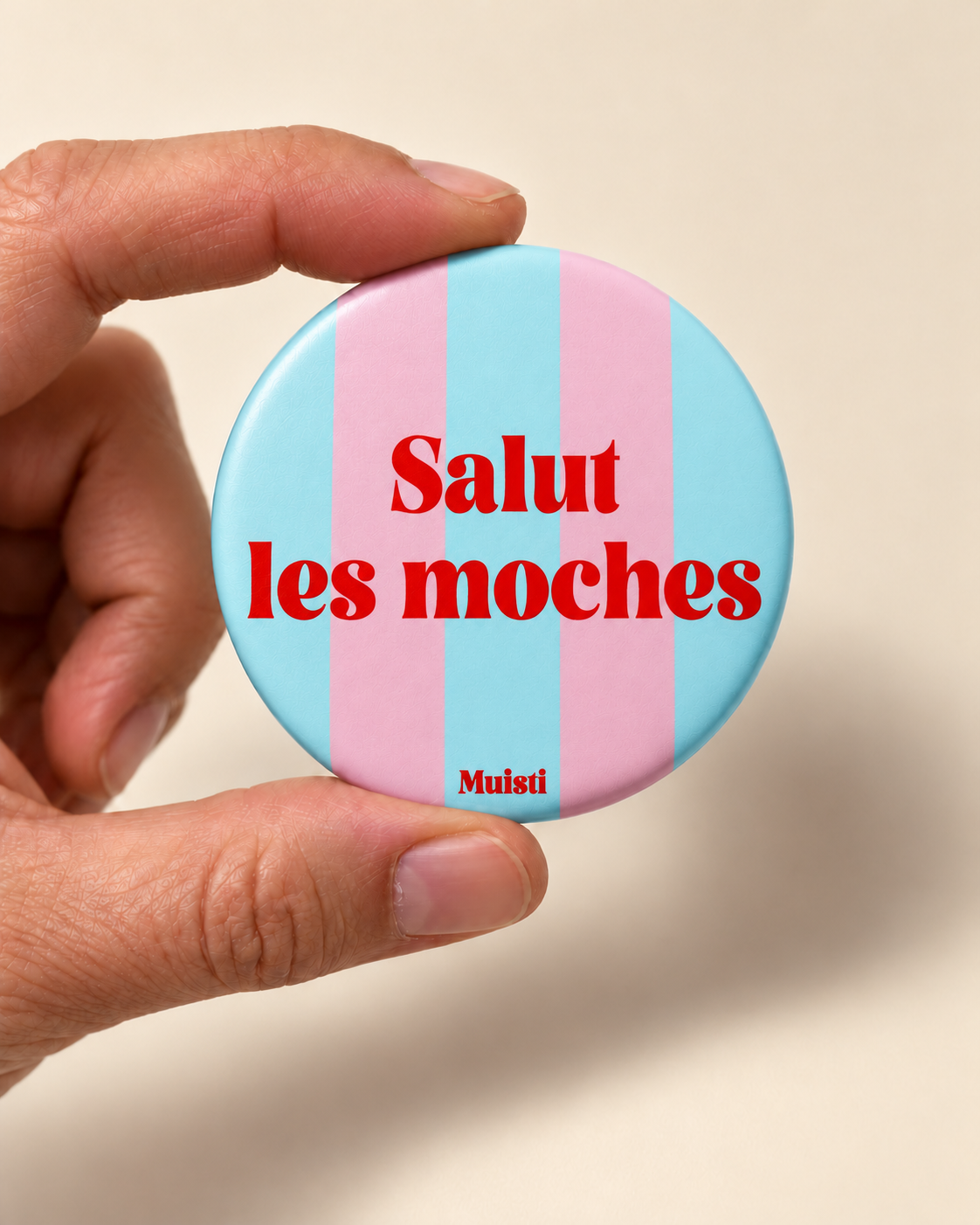 Magnet Muisti « Salut les moches » tenu entre le pouce et l’index sur fond ivoire.