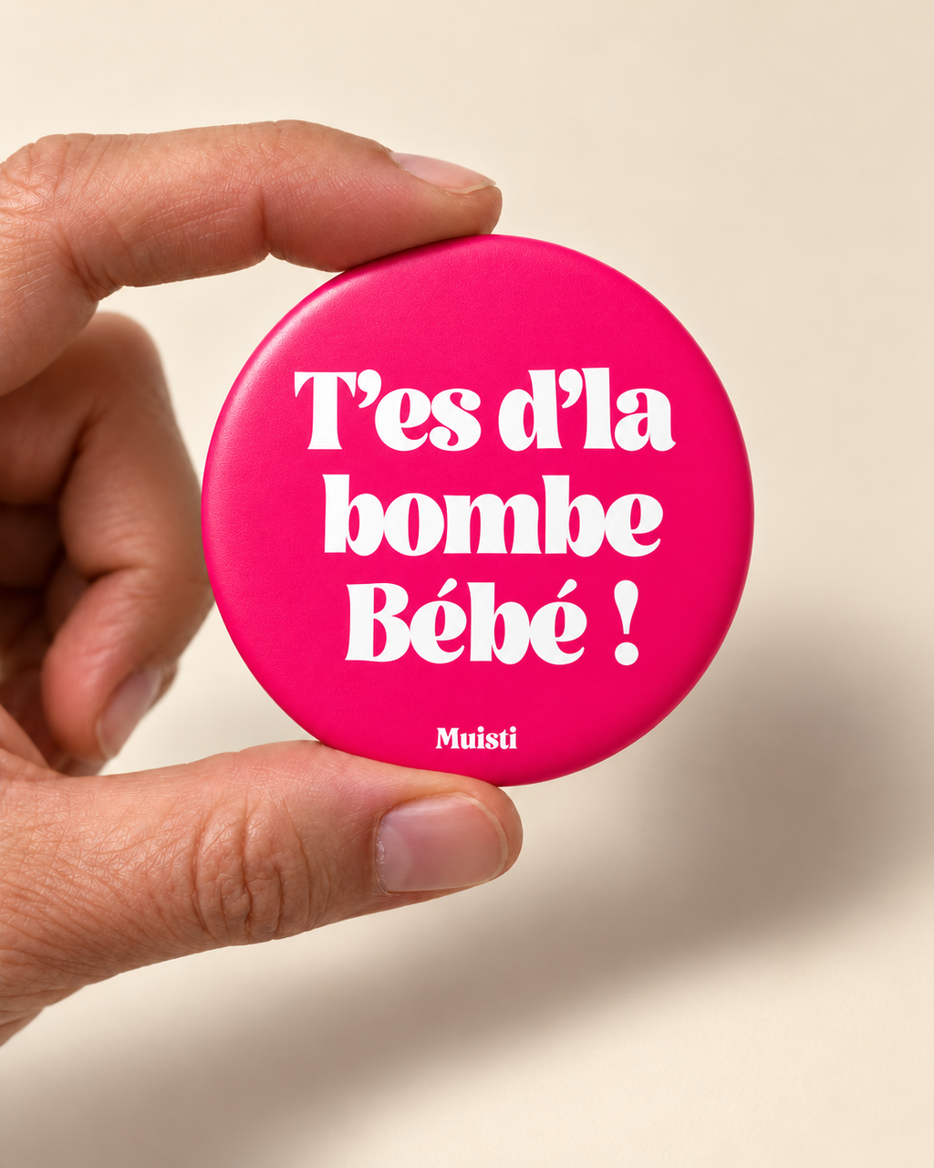 Magnet Muisti « T’es d’la bombe Bébé ! » tenu entre le pouce et l’index sur fond ivoire.