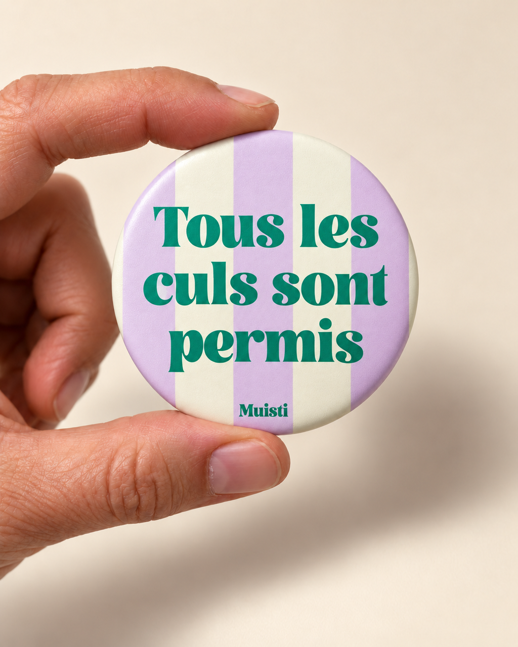 Magnet Muisti « Tous les culs sont permis » tenu entre le pouce et l’index sur fond ivoire.