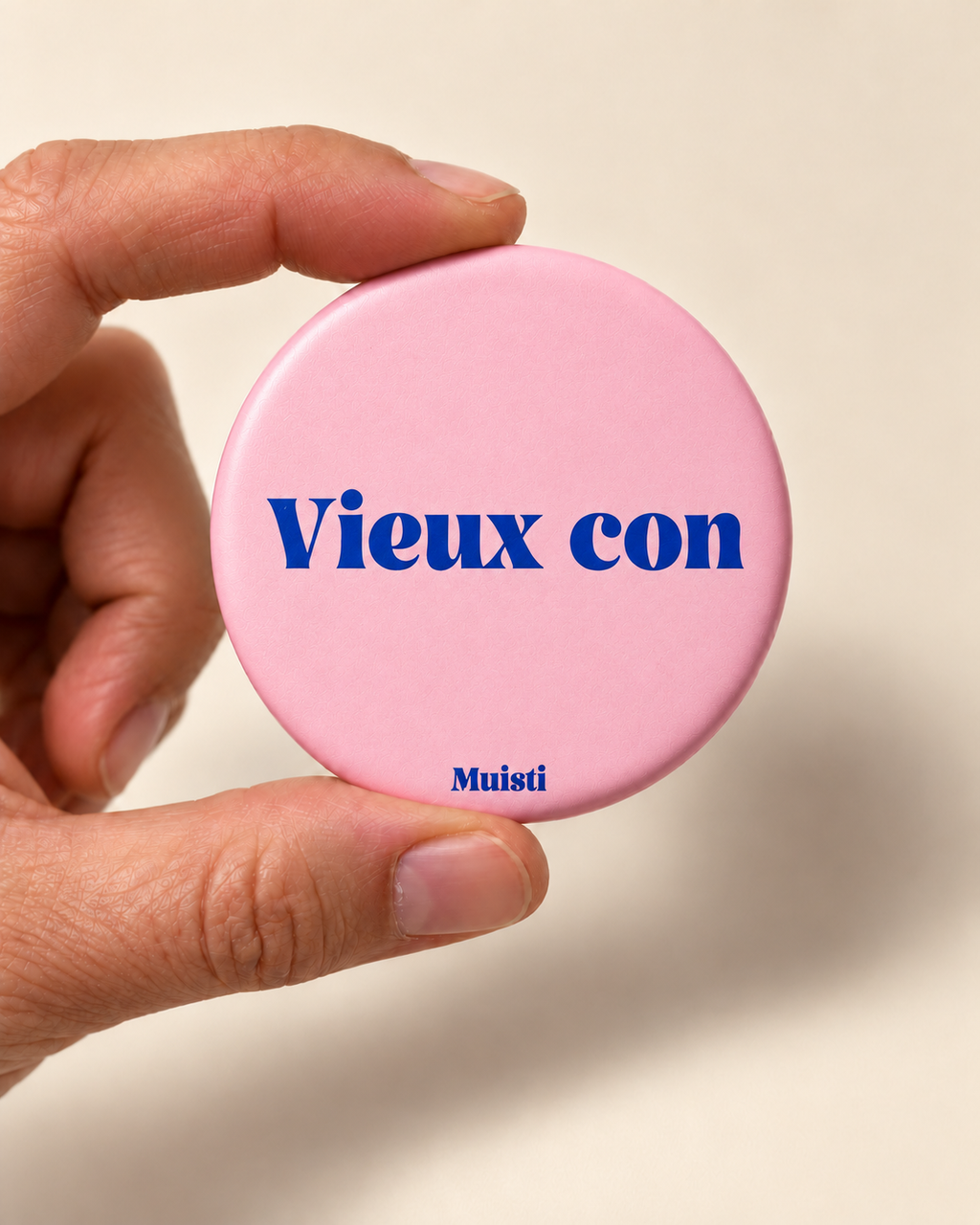 Magnet Muisti « Vieux con » tenu entre le pouce et l’index sur fond ivoire.