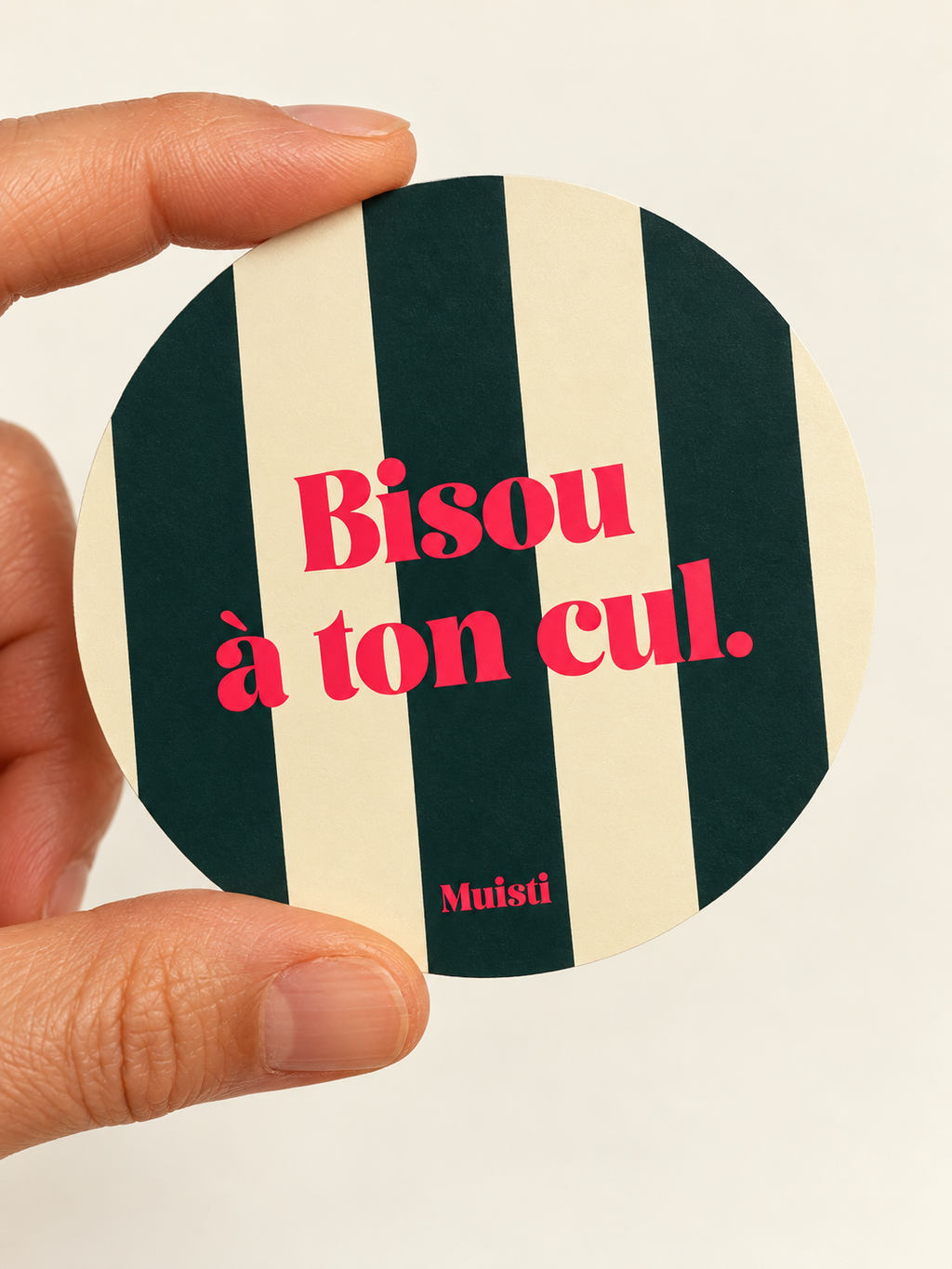 Sticker Muisti Bisou à ton cul tenu entre le pouce et l’index sur fond ivoire