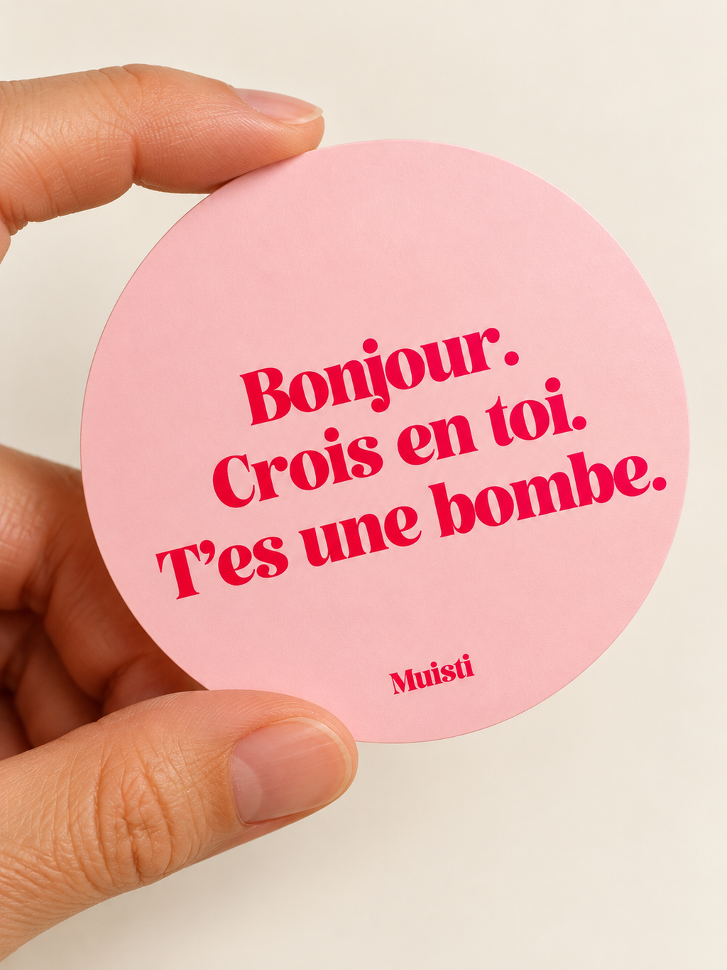 Sticker Muisti Bonjour. Crois en toi. T’es une bombe. tenu entre le pouce et l’index sur fond ivoire
