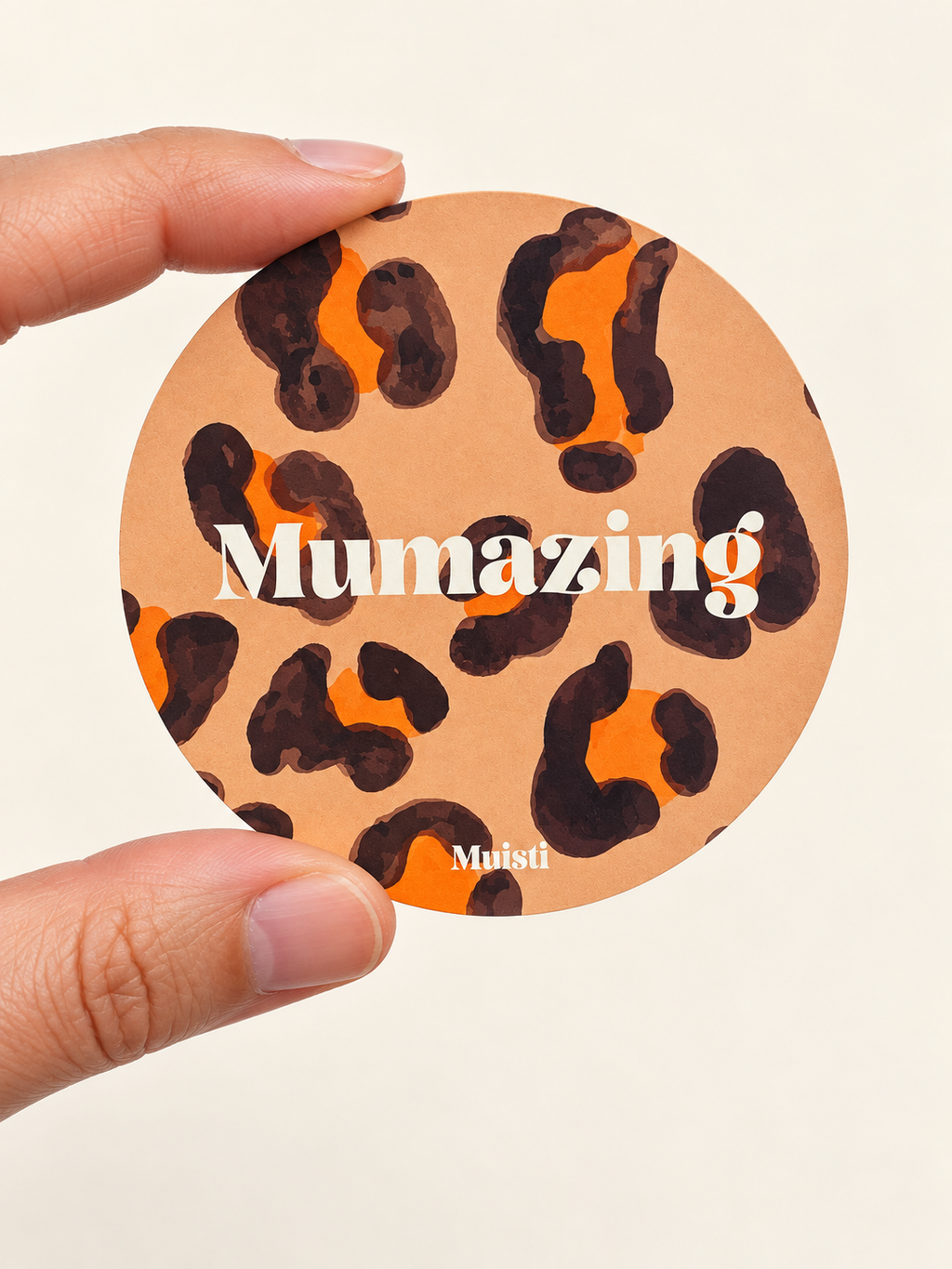 Sticker Muisti Mumazing tenu entre le pouce et l’index sur fond ivoire