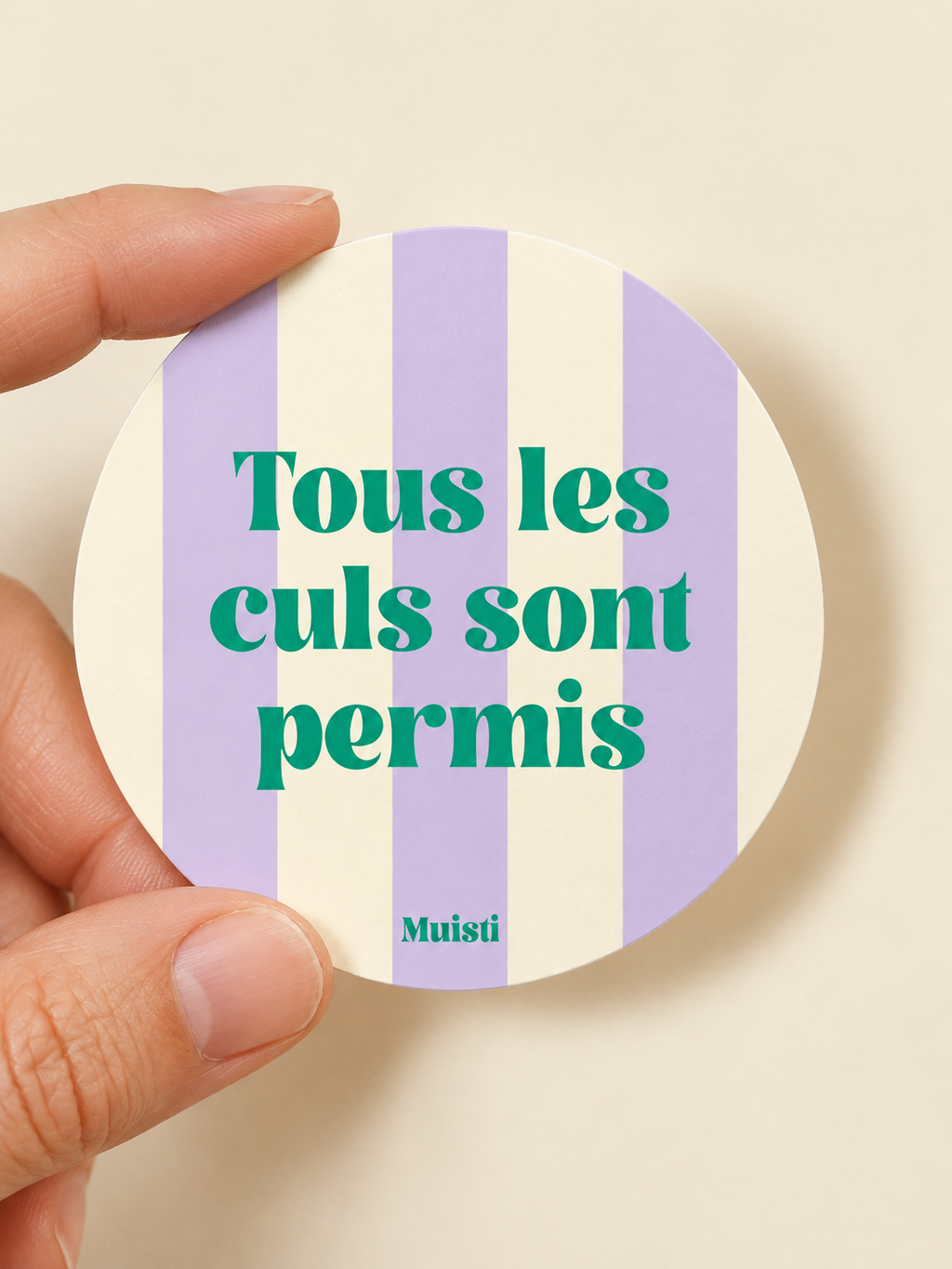 Sticker Muisti Tous les culs sont permis tenu entre le pouce et l’index sur fond ivoire